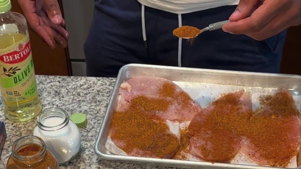 Cajun seasoning being spooned over raw chicken breasts on a parchment-lined sheet pan, with an olive oil bottle and spice jars alongside.