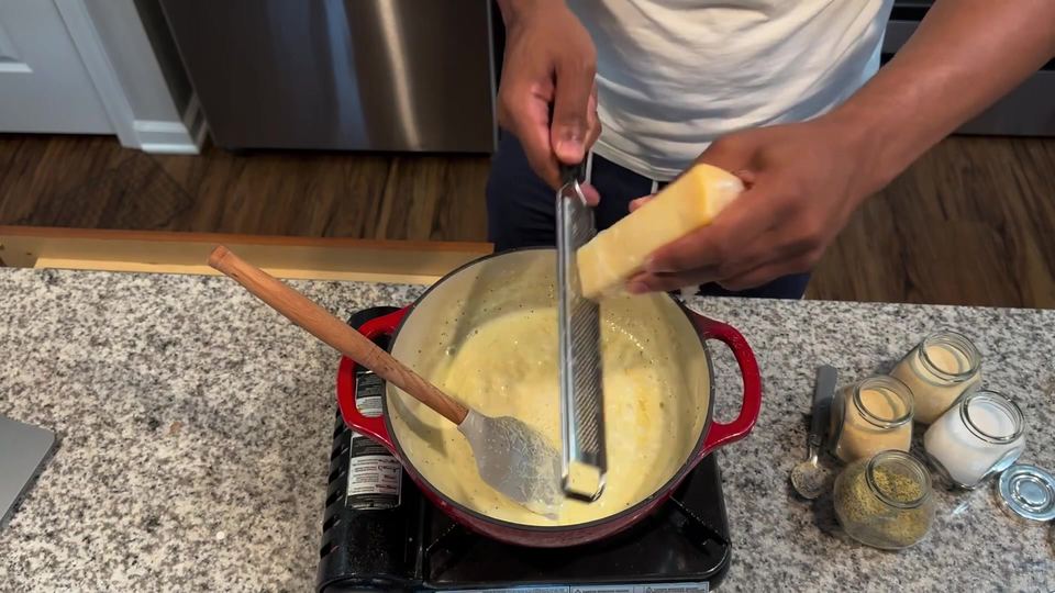 Hands grating a wedge of Parmesan on a microplane directly into pale cream sauce in a red enameled Dutch oven with a spatula resting inside.