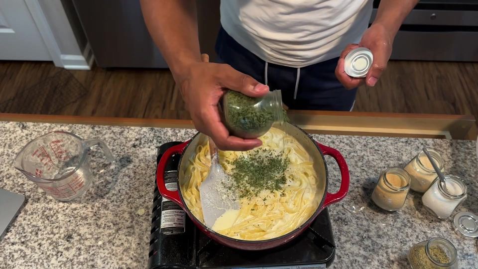 Dried parsley being sprinkled from a jar over fettuccine noodles in cream sauce in a red Dutch oven, with a fresh splash of cream visible.