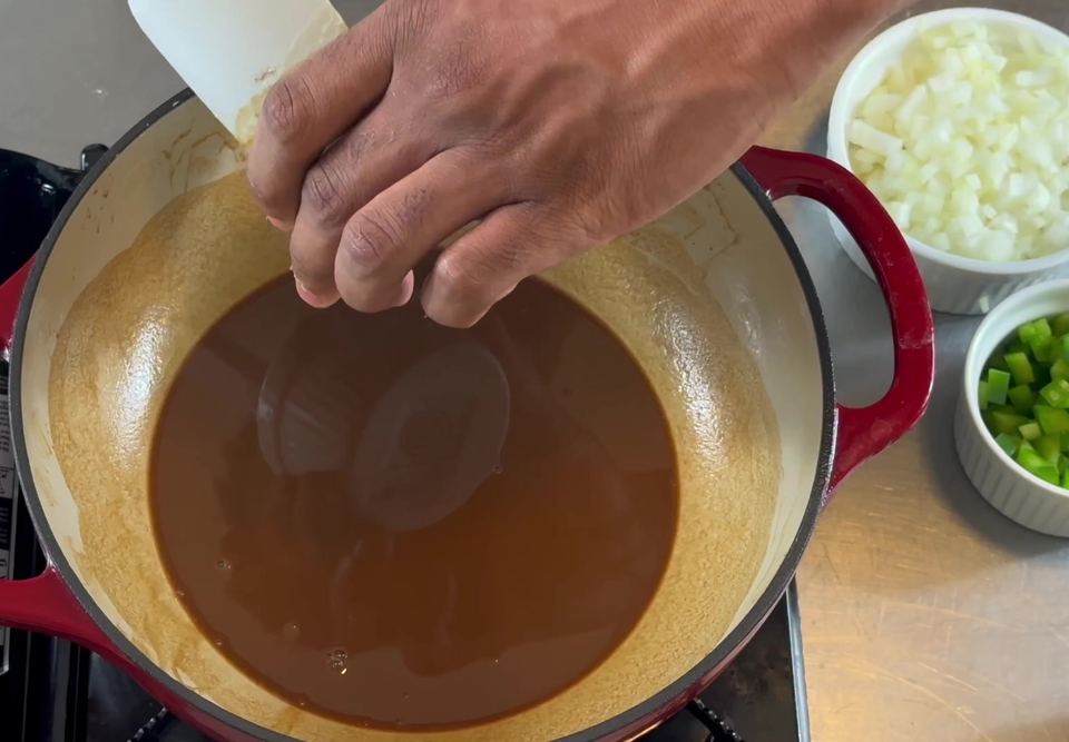 Dark brown roux in a red enameled Dutch oven with a spatula and hand above the pot, ramekins of diced onion and green bell pepper staged beside it.