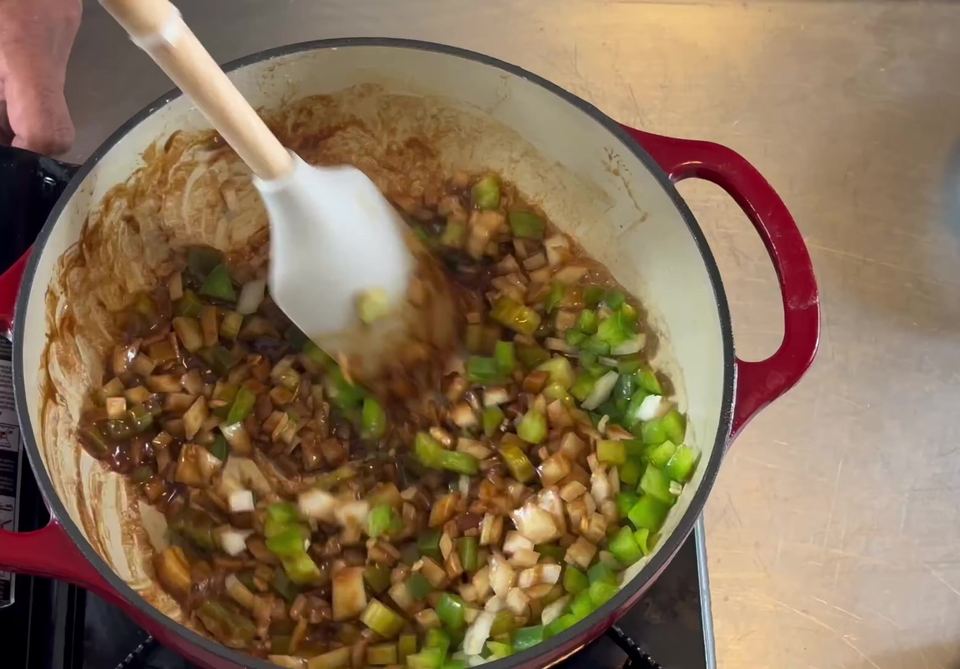 Diced celery, onion, and green bell pepper being folded into dark roux with a white spatula in a red enameled Dutch oven.
