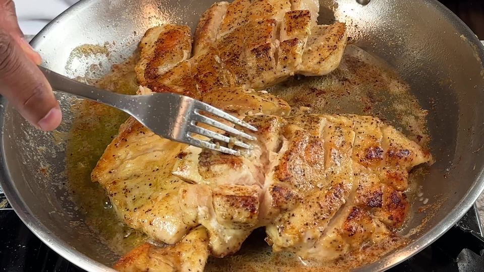 A fork pressing open a golden-brown seared chicken breast in a stainless steel skillet to show the middle is cooked through.