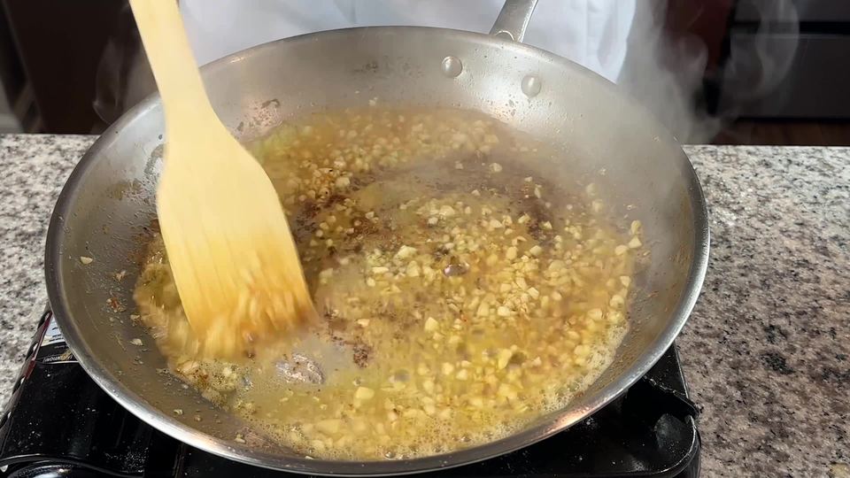 Minced garlic simmering in melted butter in a stainless steel skillet, stirred with a wooden spatula as steam rises.