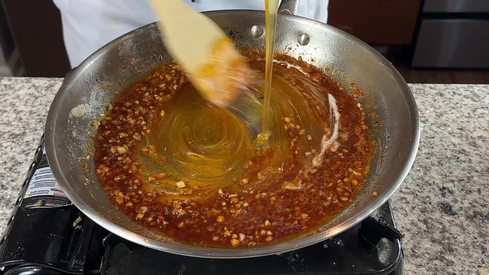 A ribbon of honey pouring into amber garlic sauce in a stainless steel skillet while a wooden spatula stirs it through.
