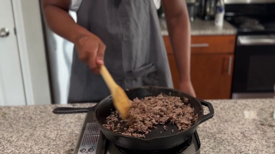 Shaved steak, browned with no pink remaining, being stirred with a wooden spatula in a cast-iron skillet on a portable burner.
