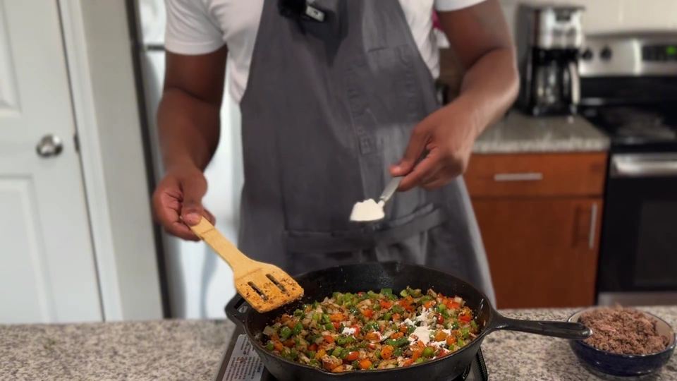 A tablespoon of flour dropped over sauteed diced bell peppers and onions in a cast-iron skillet, with a bowl of browned steak beside the pan.