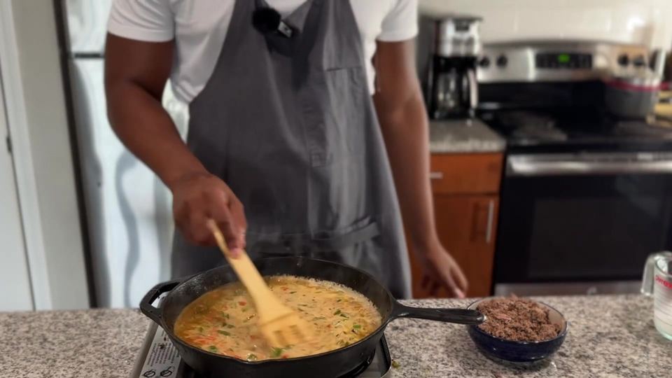 Pale golden sauce of beef broth and cream simmering over sauteed peppers and onions in a cast-iron skillet, stirred with a wooden spatula.