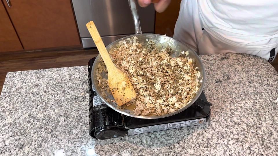 Fully cooked, crumbled ground turkey with diced onion and dried herbs in a stainless steel skillet on a portable burner, wooden spatula resting in the pan.