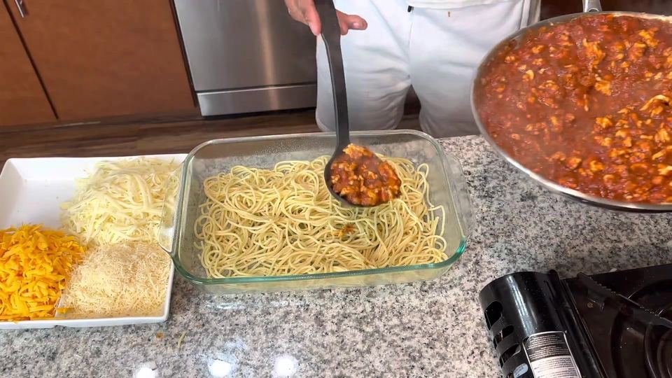 A ladle spooning red turkey sauce onto a layer of spaghetti in a glass baking dish, with trays of shredded cheddar, mozzarella and Parmesan staged beside it.