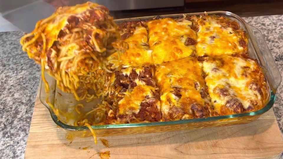 Baked cheesy spaghetti cut into squares in a glass dish on a wooden board, one portion lifted out showing the layered noodles, sauce and browned cheese.