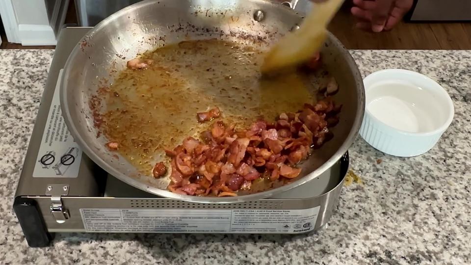 Chopped bacon frying to a deep reddish-brown in a stainless steel skillet of golden chicken drippings, stirred with a wooden spatula.