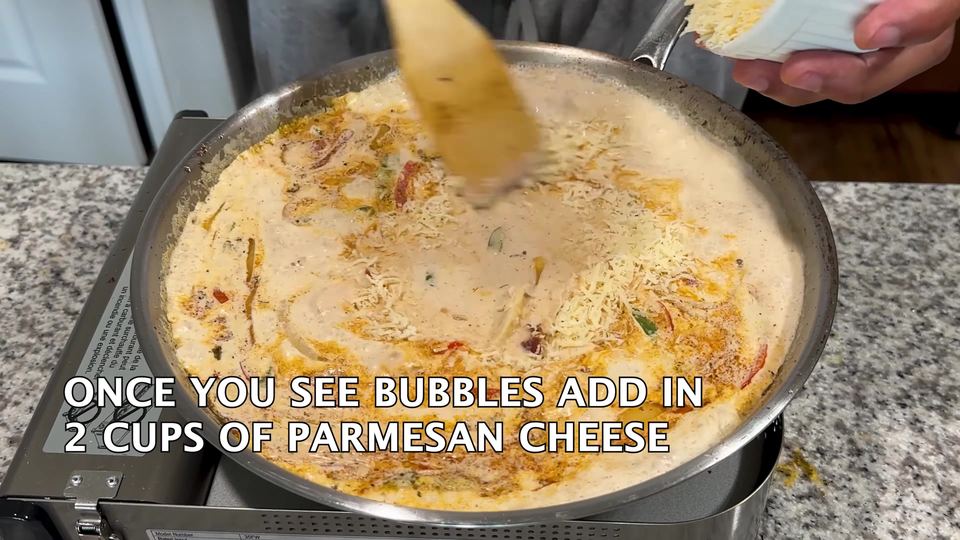 Pale cream sauce bubbling in a large stainless skillet with peppers and onions, as shredded Parmesan cheese is sprinkled over the top.