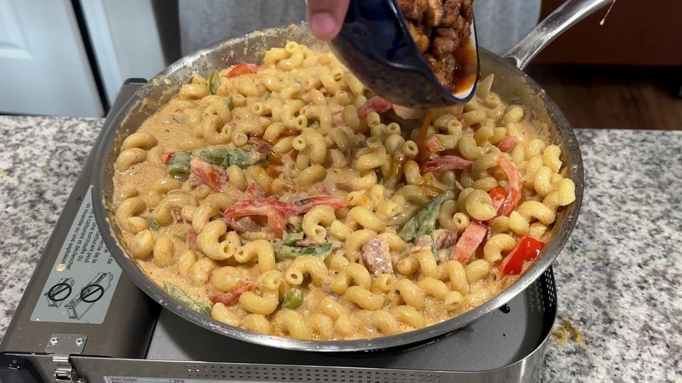 Cavatappi pasta coated in a creamy orange sauce with red and green bell pepper strips, as cooked chicken is poured in from a blue bowl.