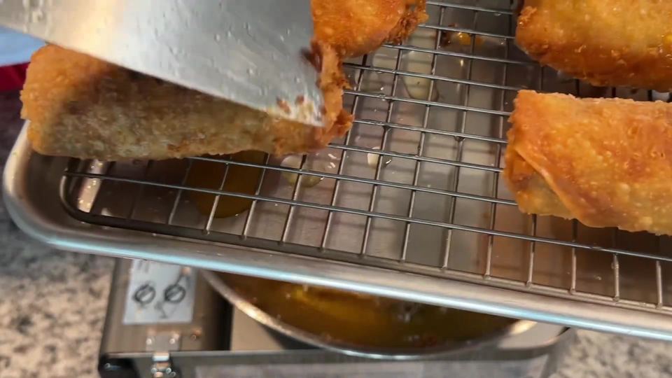 A dark golden egg roll being lifted onto a wire cooling rack with a metal strainer, more crispy fried rolls resting alongside and the oil pot below.
