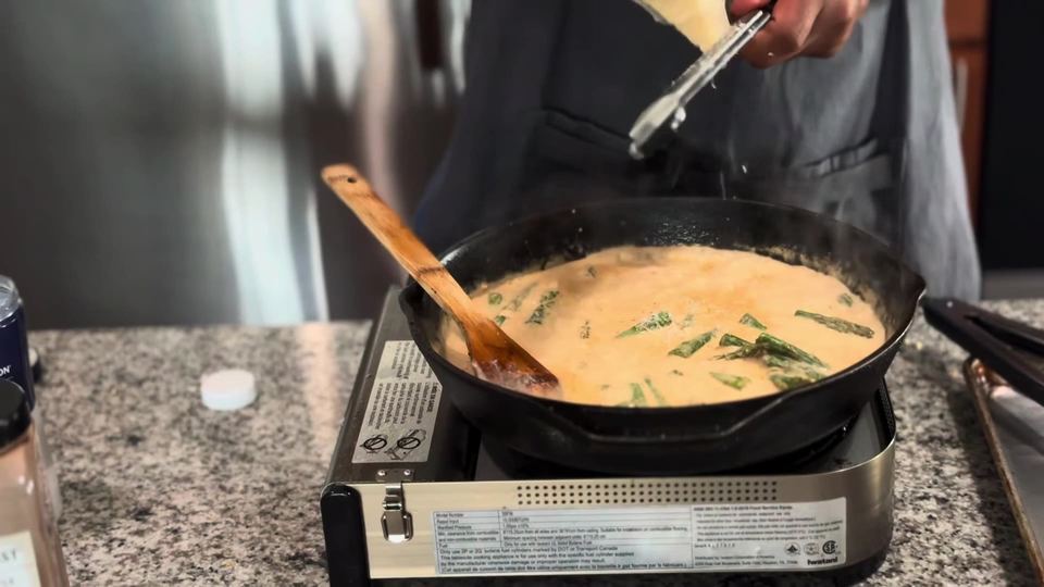 Parmesan being grated into a bubbling cream sauce with asparagus pieces in a cast-iron skillet, a wooden spoon resting in the pan.