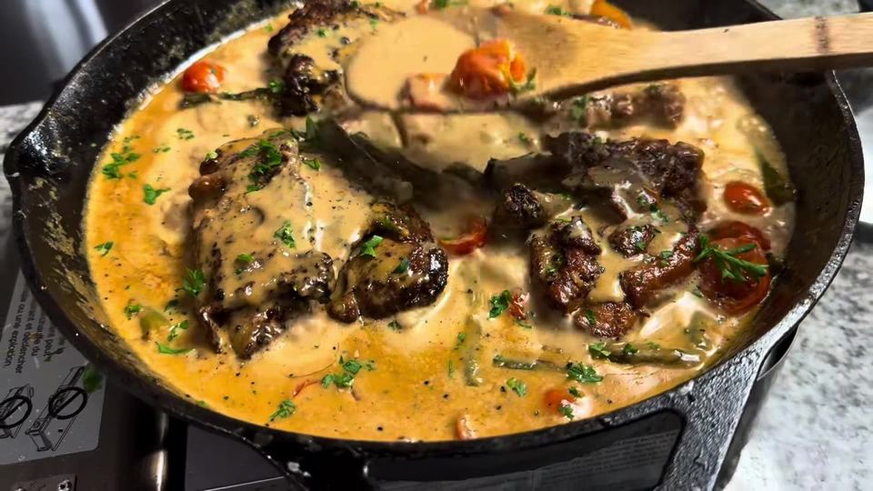 Wooden spoon basting creamy Cajun Parmesan sauce over chicken thighs, asparagus, and cherry tomatoes in a cast-iron skillet.