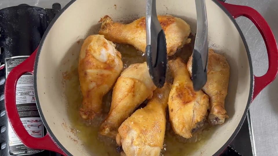 Golden-browned chicken drumsticks frying in shallow oil in a red Dutch oven, being turned with tongs.