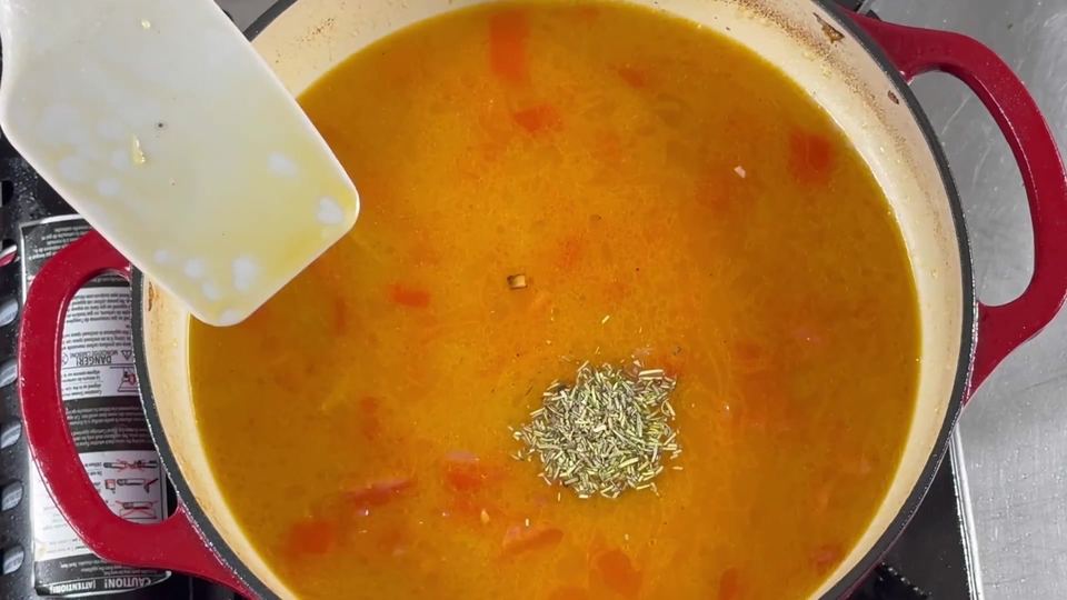 Bright orange tomato-and-broth base in a red Dutch oven with a small pile of dried herbs added in the center.