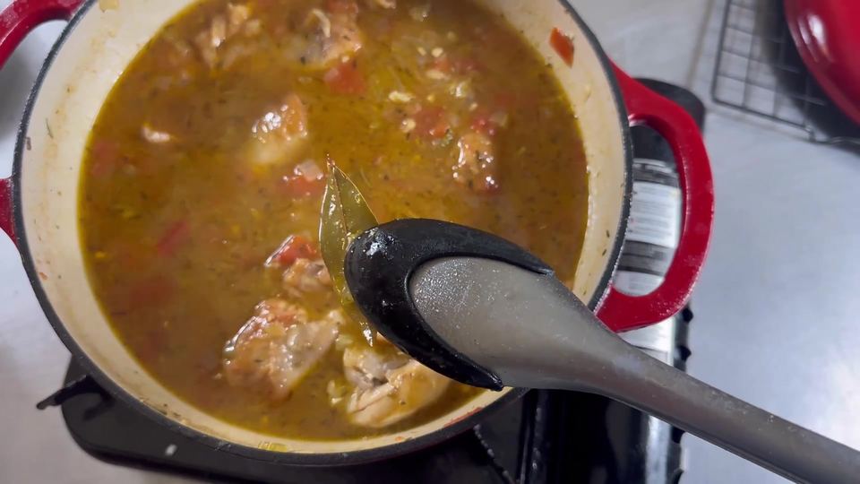 Finished Creole stew chicken in a red Dutch oven with dark rich broth and tender chicken; a spoon lifts out the bay leaf.