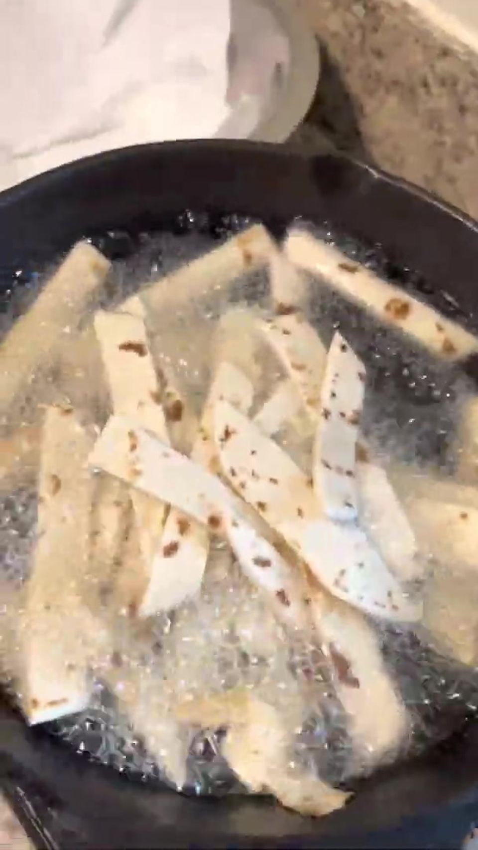 Pale tortilla strips floating in vigorously bubbling hot oil in a dark skillet.