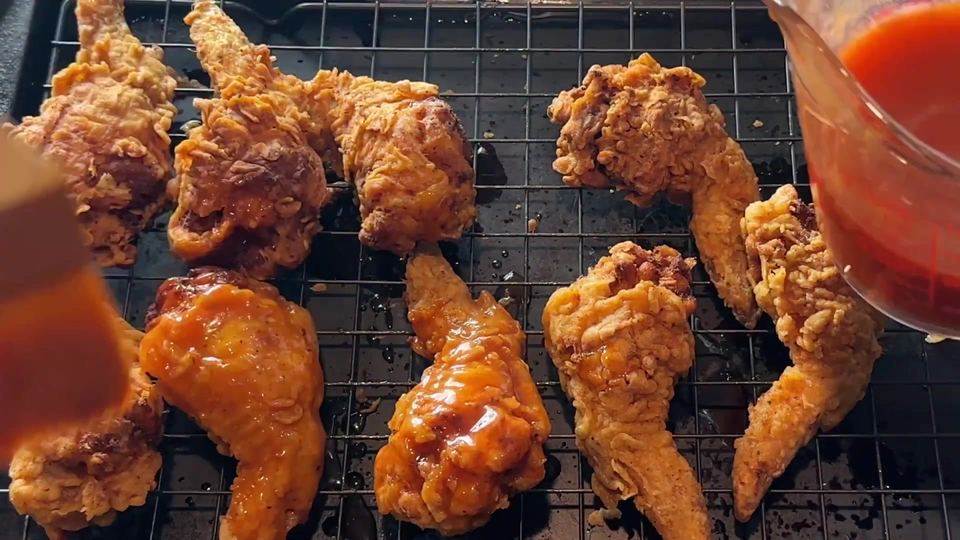 Hot honey sauce pouring from a glass measuring cup over deep golden fried stuffed wings resting on a black cooling rack.