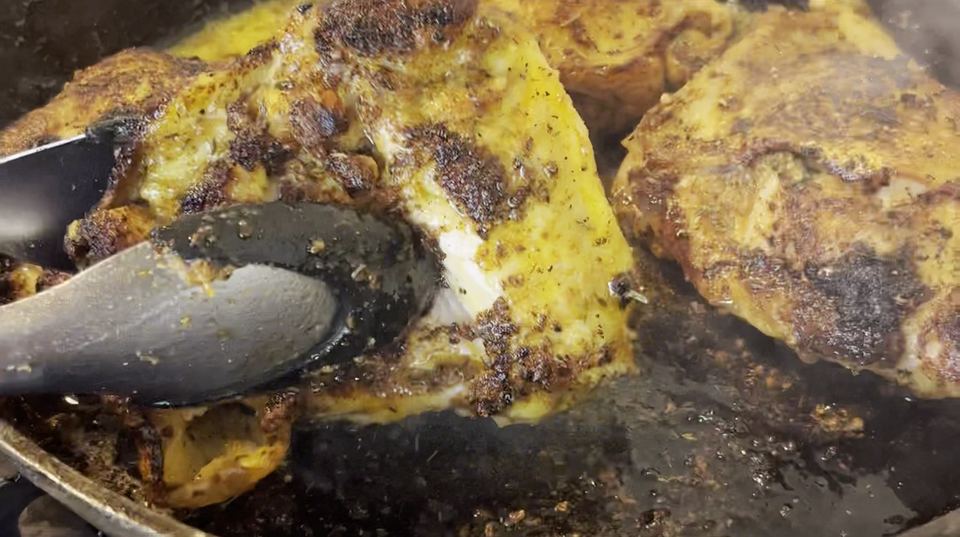 Seasoned chicken thighs searing in a cast-iron skillet, one lifted with tongs to reveal a deeply browned crust.