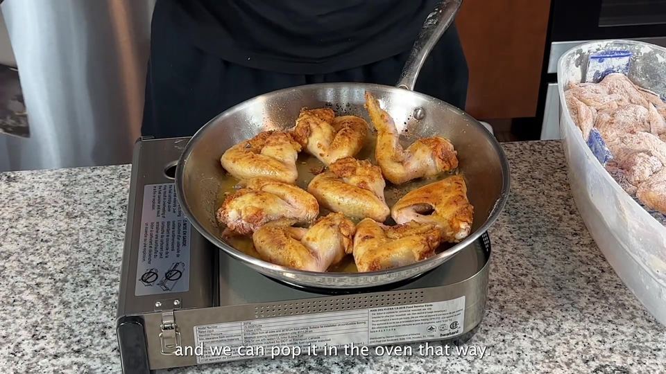 Chicken wings frying golden brown in a stainless steel skillet on a portable burner, with a bin of flour-dredged raw wings waiting beside the stove.
