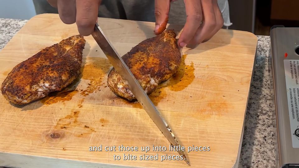 Two chili-seasoned, browned chicken breasts on a wooden cutting board, one being sliced with a long knife to show the cooked interior.