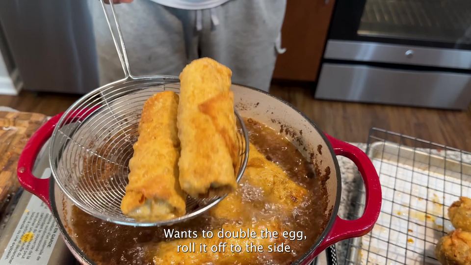 Two golden-brown fried egg rolls lifted on a wire spider strainer above a red Dutch oven of bubbling frying oil.