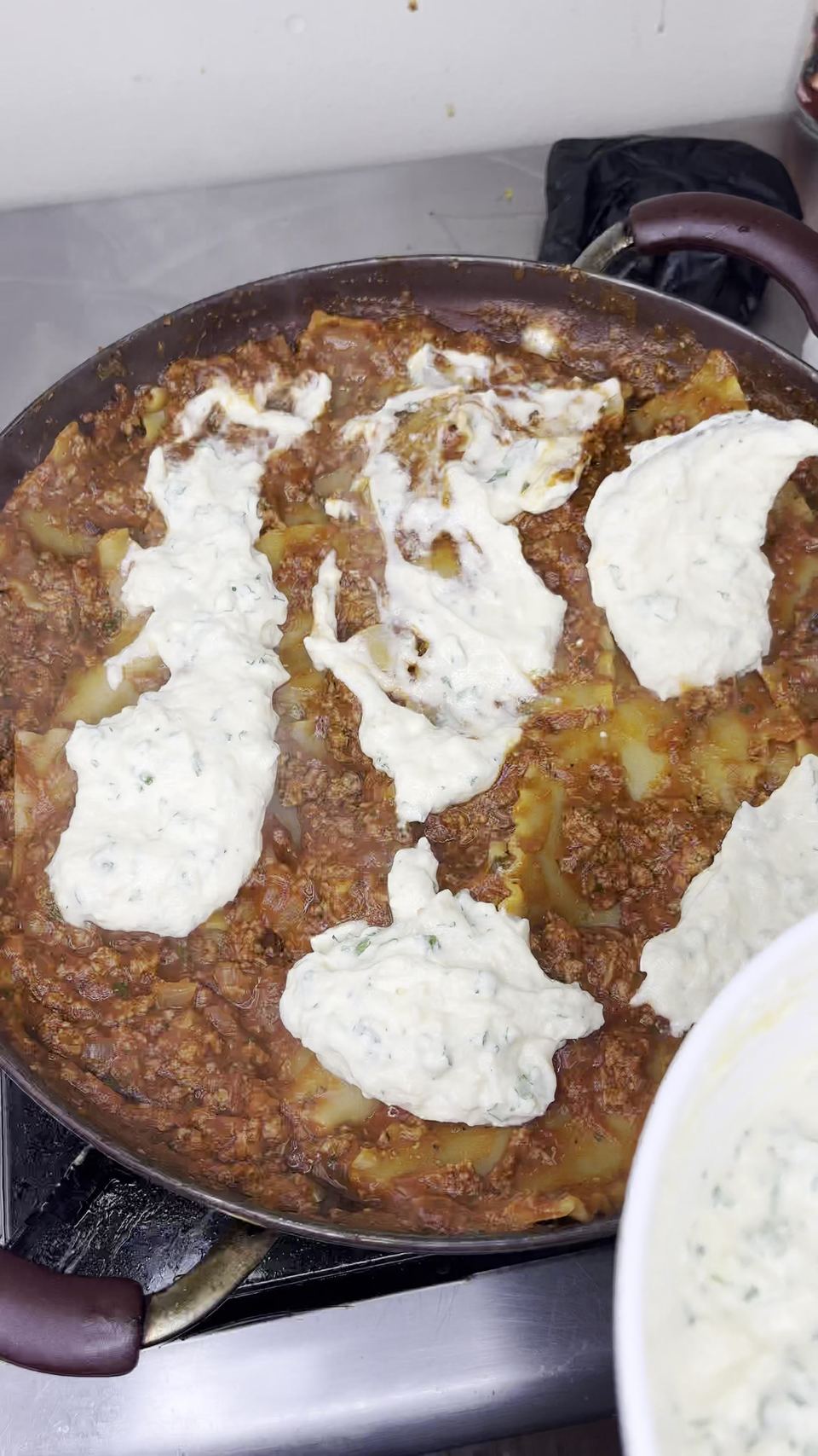 Skillet of meat sauce and noodles topped with large dollops of parsley-flecked ricotta-egg mixture; a bowl of the remaining ricotta mix sits at the lower right.